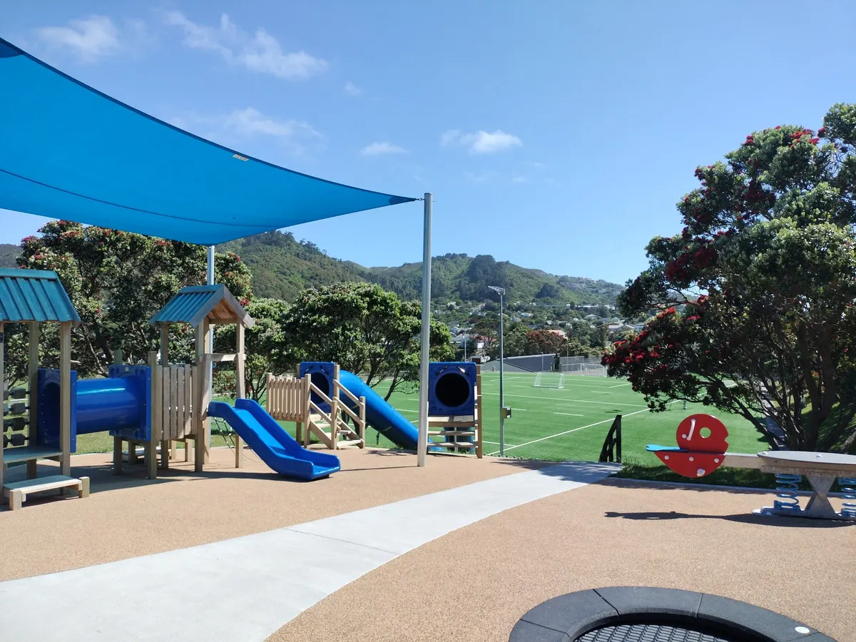Blue slides, tower and in-ground trampoline under a shade sail beside the football turf at Nairnville Park playground