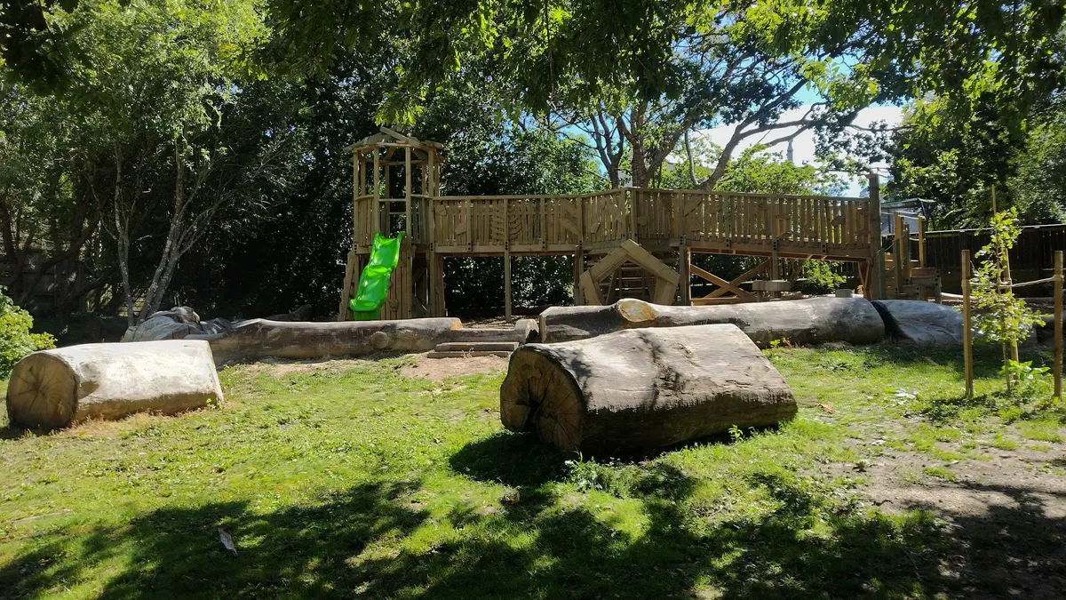 Timber fort with green slide and log play under trees at Whitby Walkway playground, Porirua