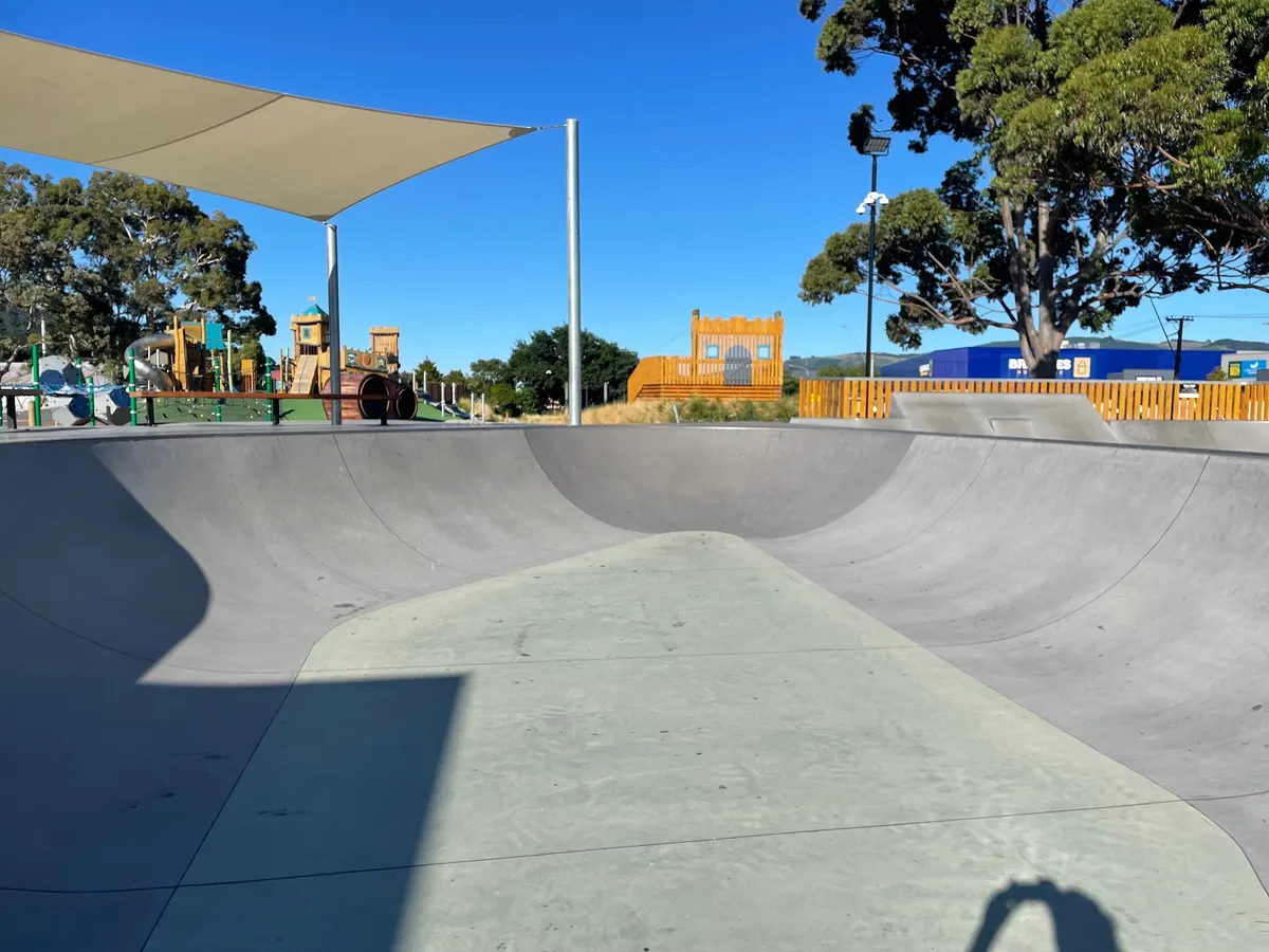 Concrete flow bowl under a shade sail at Maidstone Skatepark, Upper Hutt, with the castle playground behind