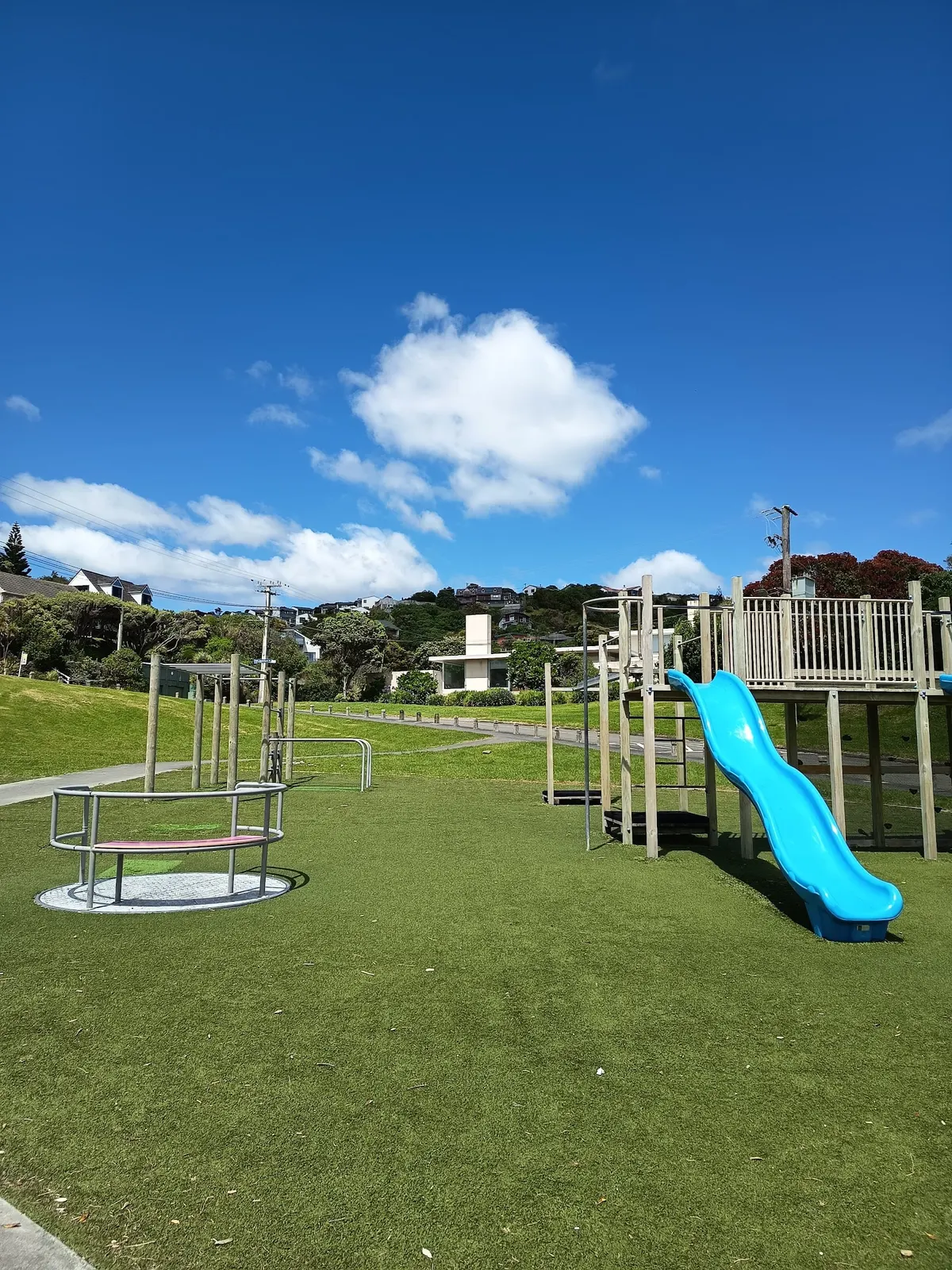 Inground carousel and blue wavy slide on artificial grass at Arnold Park playground, Tītahi Bay