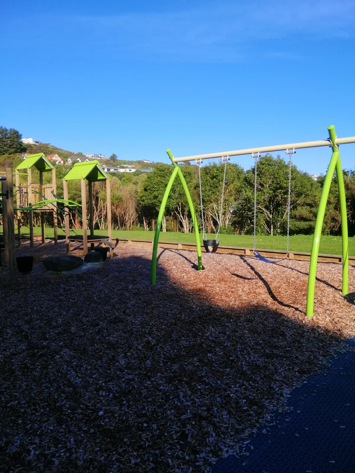Swing frame and green-roofed play tower on bark at Moray Playground, Papakōwhai, Porirua