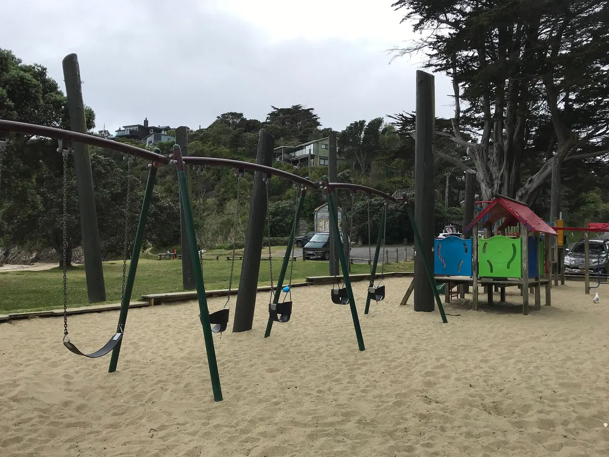 Row of swings with infant seats on sand beside timber play kit at Children's Playground, Oneroa, Waiheke