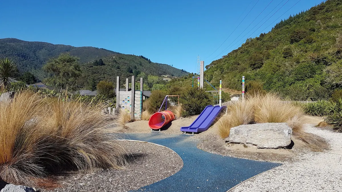 Red tunnel and purple slide on grassed mounds with bush hills behind Hockey Reserve playground, Nelson