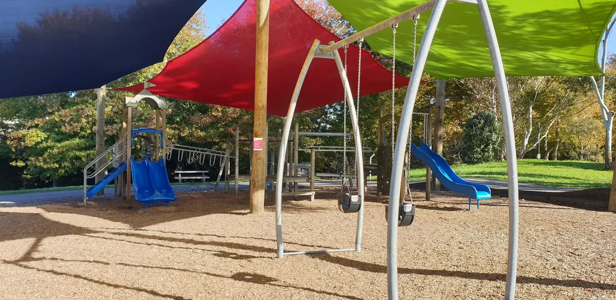 Swings and a blue slide under coloured shade sails at Shepherds Park playground, Beach Haven