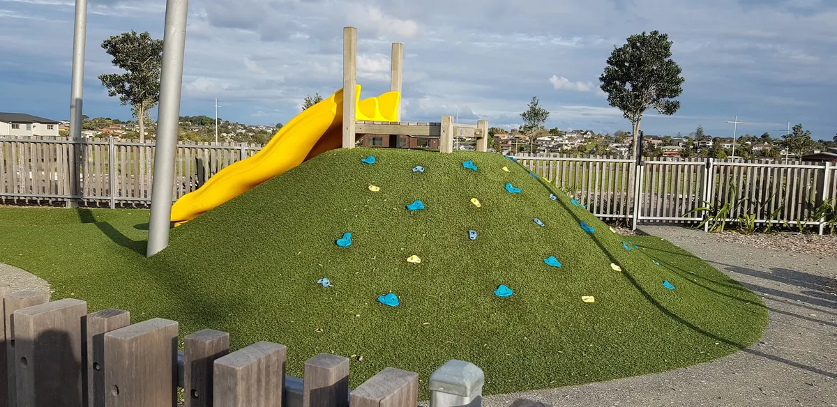 Grass climbing hill with holds and a yellow slide at Ridge Park playground, Long Bay