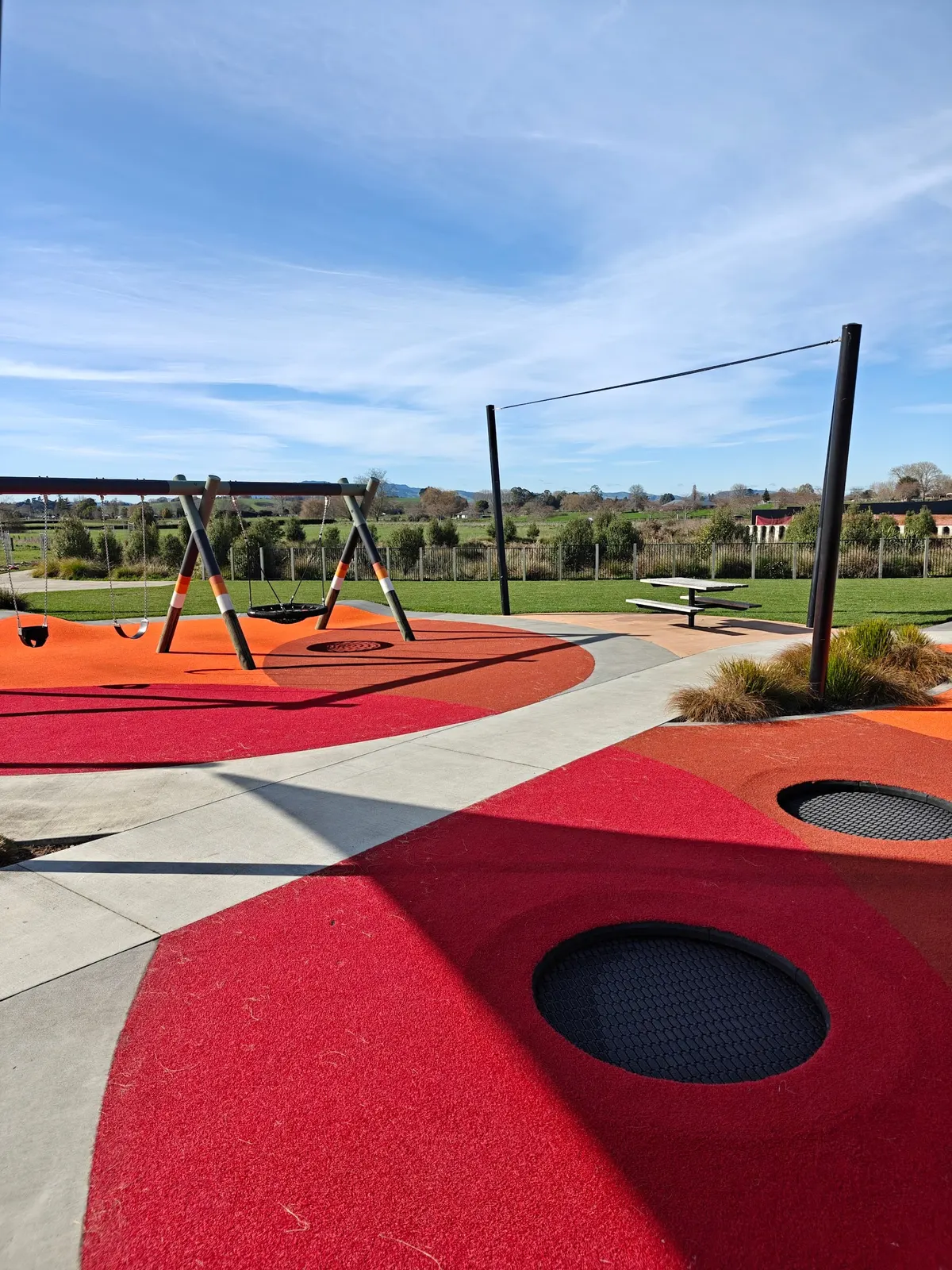 In-ground trampolines and basket swing on red rubber surfacing at Swarbrick Drive Reserve playground, Te Awamutu