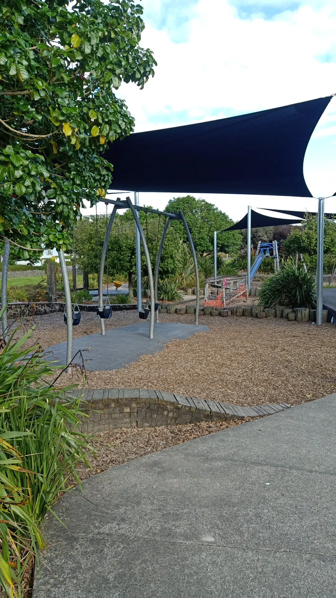 Infant swings and navy shade sails over the bark play area at Hobsonville Point playground