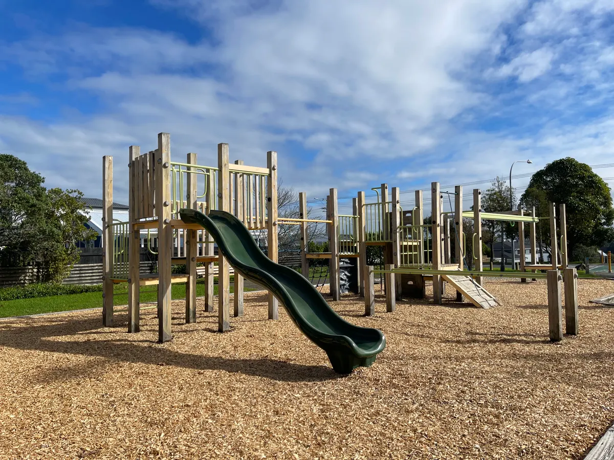 Wooden multi-play tower with curved green slide on bark at Bayswater playground, Auckland