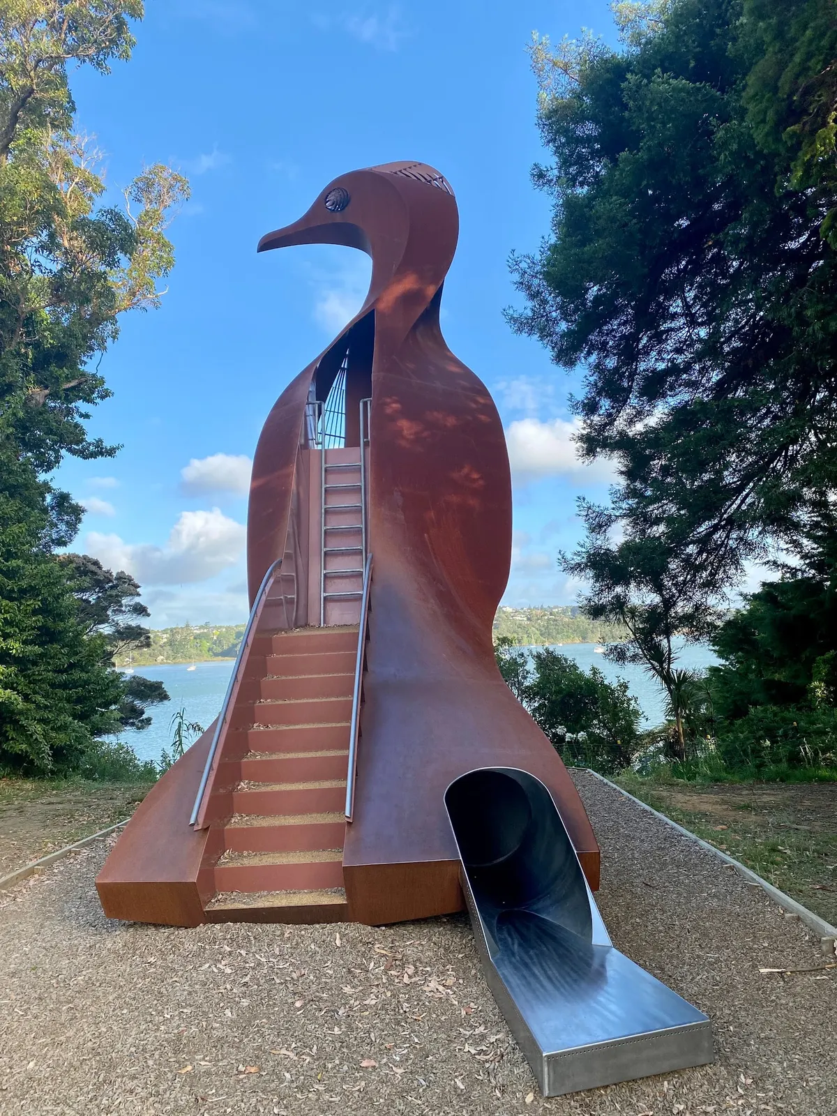 Ten metre rust-coloured pied shag sculpture with steps and a metal slide at Harrier Point Park playground, Hobsonville