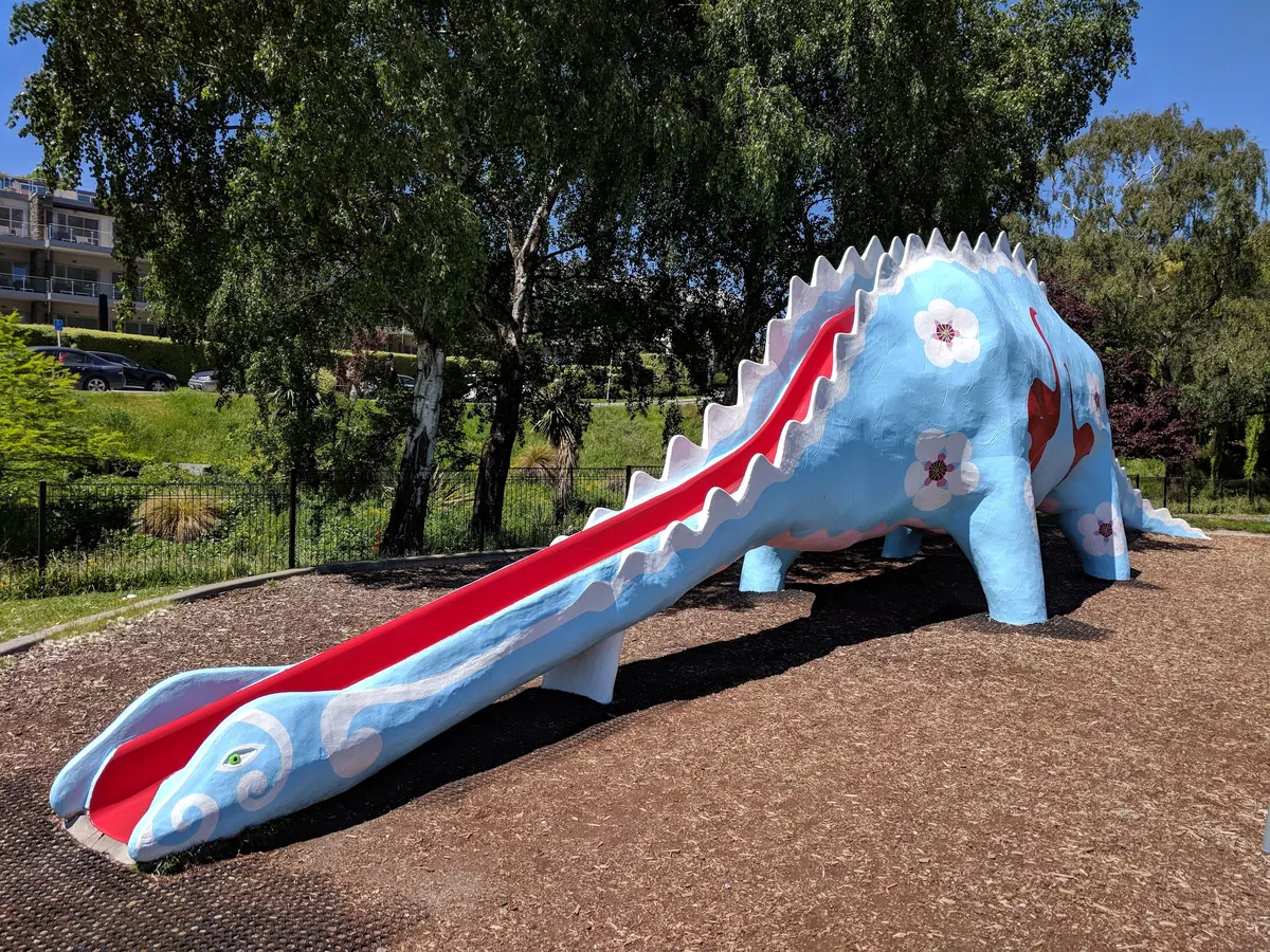 Blue dinosaur slide with flower and moa motifs on bark at Wānaka Dinosaur Playground